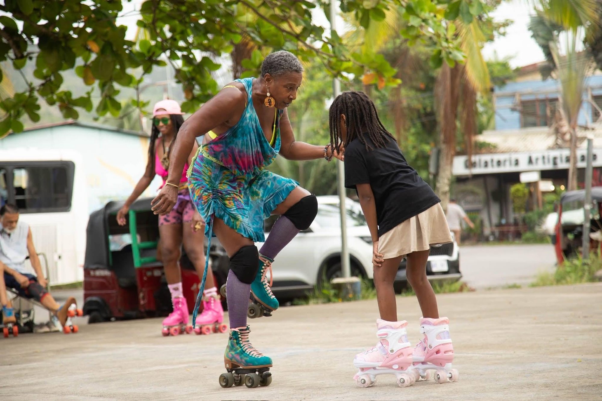 A coach in a tie-dye dress guiding a young girl on her first roller skates in Puerto Viejo
